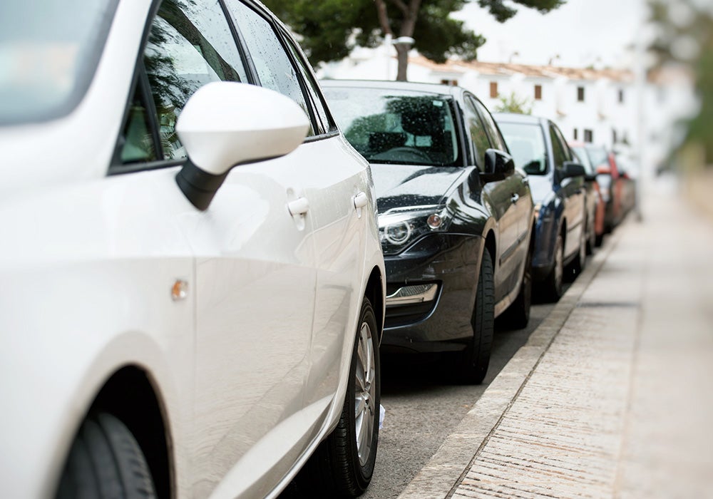 Line of parked cars on a city street, with a white vehicle in the foreground and a black car behind it.