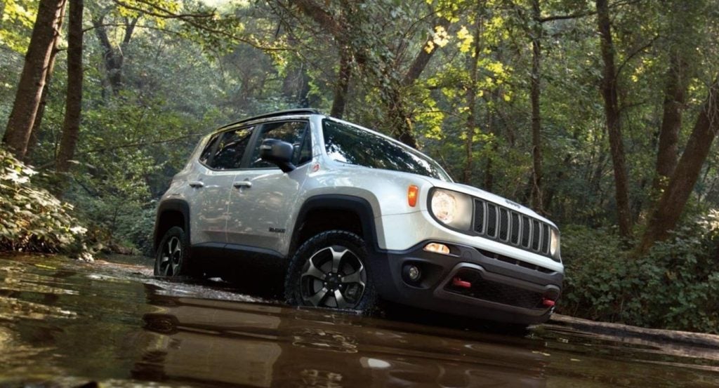 A white Jeep Renegade fords a shallow stream in a lush, green forest.