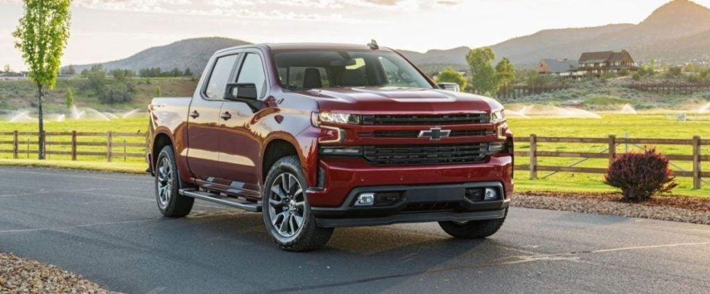 Red Chevy Silverado truck parked on asphalt with a fenced green field, mountains, and a large house in the background.