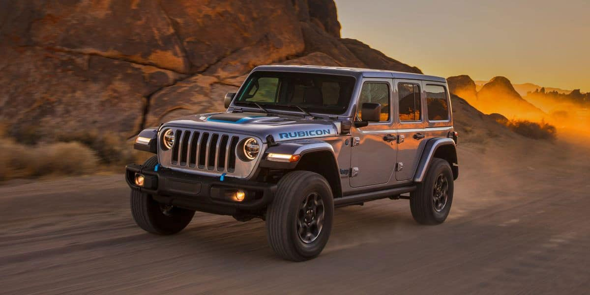 A silver Jeep Rubicon with blue accents drives off-road in a sandy desert under an orange sunset.