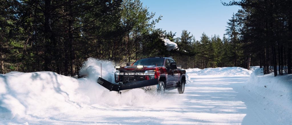 Red Chevrolet pickup truck with a "BOSS" snowplow clearing a snowy forest road, kicking up snow.