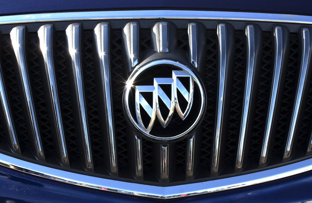 Close-up of a silver Buick logo on a dark blue vehicle's front grille, with sunlight glinting off the emblem.