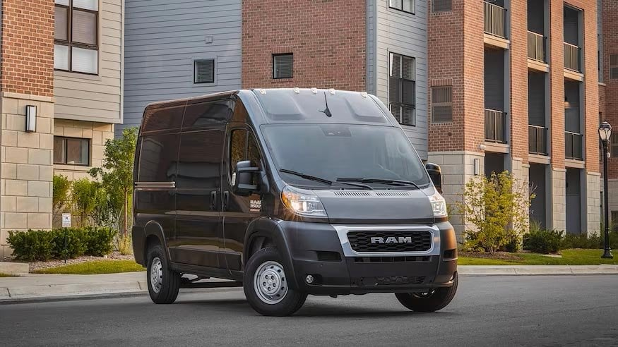 A gray Ram ProMaster cargo van parked on a street in front of apartment buildings.
