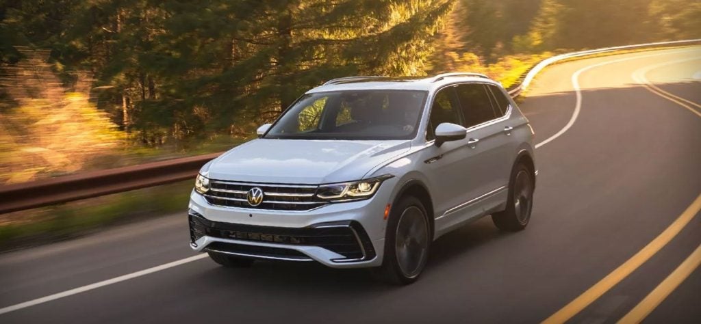 White Volkswagen Tiguan SUV driving on a winding road with trees and a luminous sunset background.