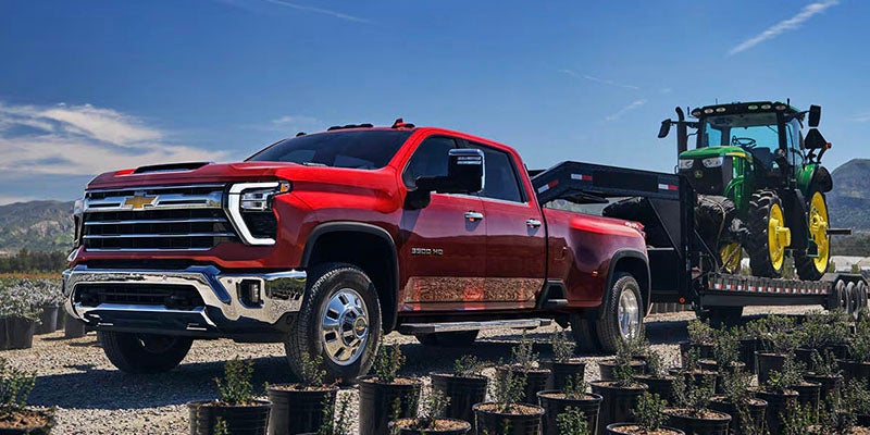 A red Chevrolet Silverado 3500 HD truck with a green John Deere tractor on a trailer behind it.