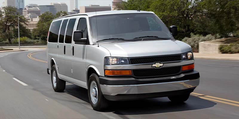 Silver Chevrolet Express passenger van driving down a curved road with a city skyline in the background.