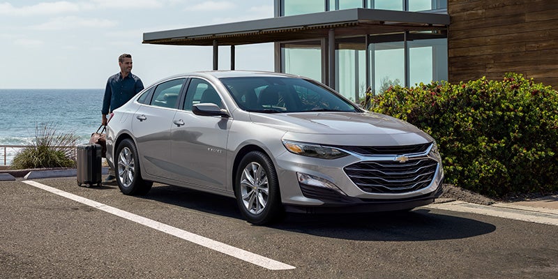 A man with luggage next to a silver Chevrolet Malibu parked by a building overlooking the ocean.