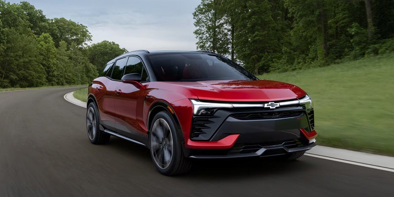 A red Chevrolet Blazer electric vehicle drives on a winding road with green trees in the background.