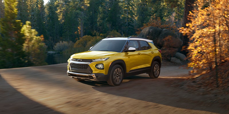 Yellow Chevrolet Trax driving on a dirt road through a forest with a pond and autumn trees.