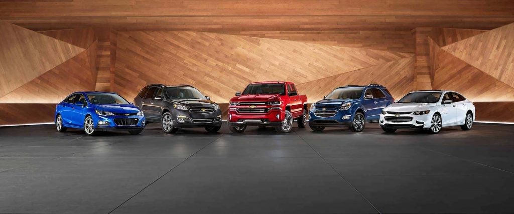 Five Chevrolet vehicles, including a blue car, black SUV, red truck, blue SUV, and white car, on a dark floor in front of a modern wood-paneled wall.