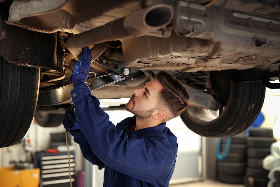 A male auto mechanic in blue overalls and gloves working under the raised chassis of a car.