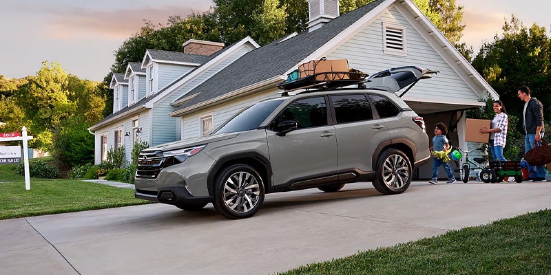 Light gray SUV with opened trunk and roof rack, parked in driveway of a blue house, family unpacking.