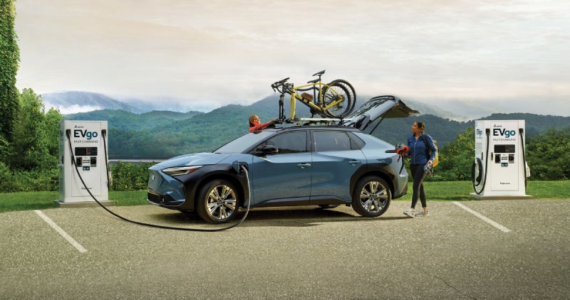 Two people charge a blue SUV with a bicycle on the roof rack at an EVgo station with mountains in the background.