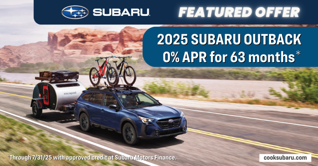 A blue 2025 Subaru Outback on a desert road, towing a small trailer, with two bikes on its roof rack.