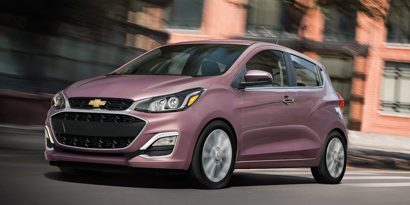 A pink/purple Chevrolet Spark hatchback driving on a city street with motion blur in the background.