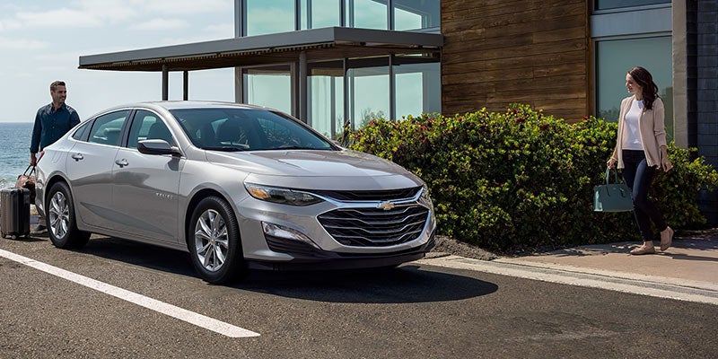Silver Chevrolet Malibu parked on a paved driveway with a man and woman approaching a modern home.