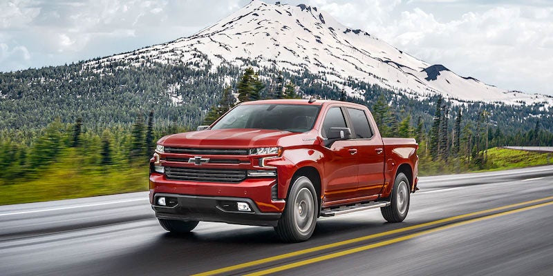 Red Chevrolet Silverado pickup truck driving on a highway with a snowy mountain and evergreen trees in the background.