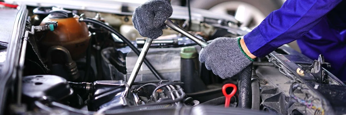 Mechanic in overalls pointing at a car engine while holding a pen and clipboard.
