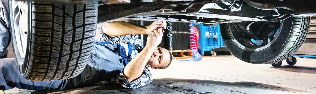 Auto mechanic in blue uniform and gloves making repairs underneath a car, illuminated by a work light.