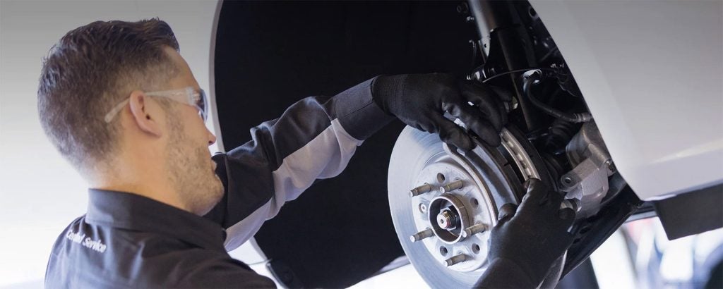 An automotive technician in safety glasses and gloves works on a car's disk brake assembly.