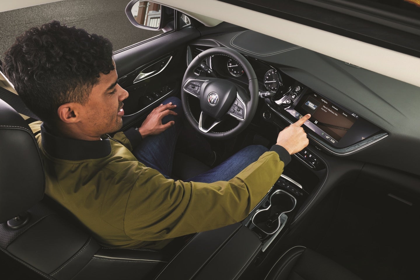 A man in a green jacket sits in a car, looking right and touching the infotainment screen.