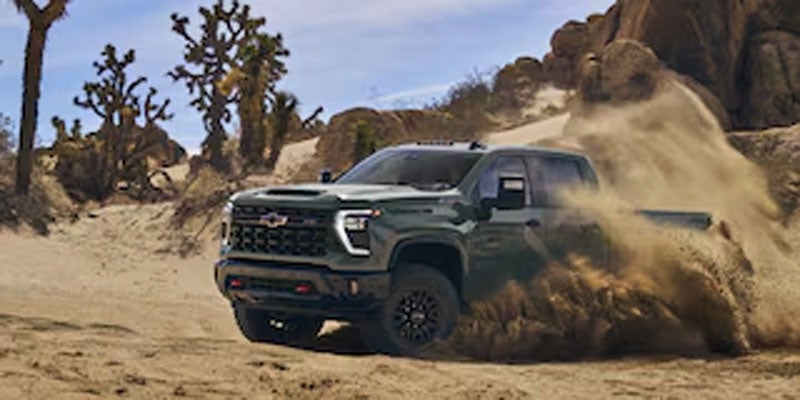 Chevrolet Silverado 2500HD kicks up a cloud of dust while driving through a winding desert path with Joshua trees and rocks.