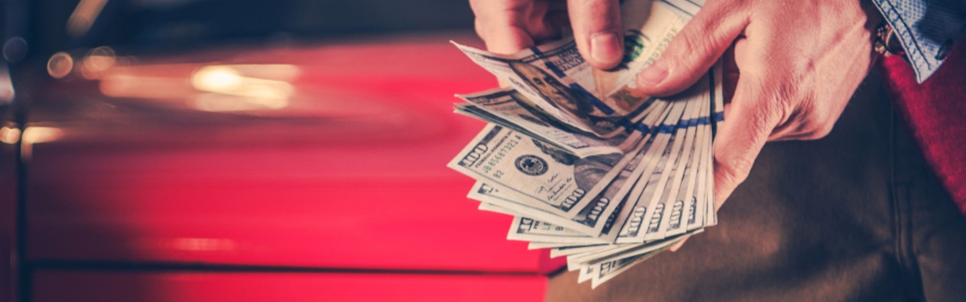 Two hands count a fan of hundred-dollar bills against a blurred red background.