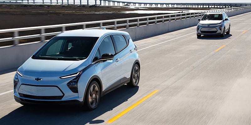 Two white Chevrolet Bolt EVs drive on a multi-lane highway over a body of water with a distant bridge.