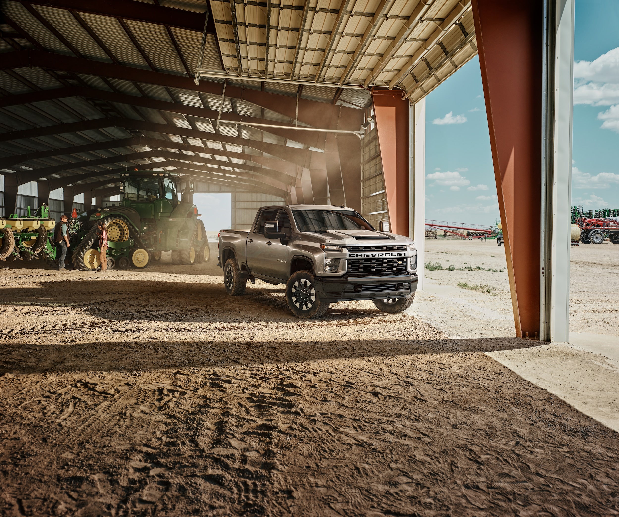 A silver Chevrolet Silverado HD truck in a barn, open to a dusty farm field with a blue sky.