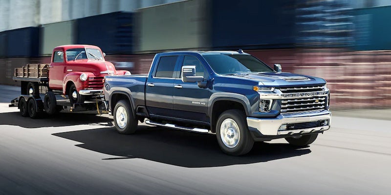 A blue Chevrolet Silverado HD truck pulling a red vintage Chevy truck on a flatbed trailer down a road.