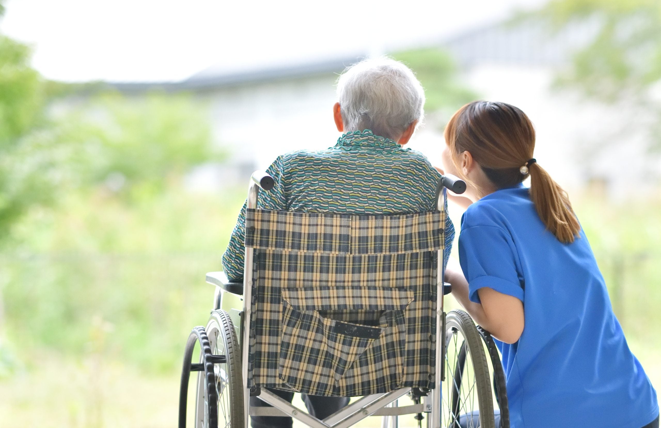 A caregiver is crouched down next to a person in a wheelchair, and they are looking out into nature together.