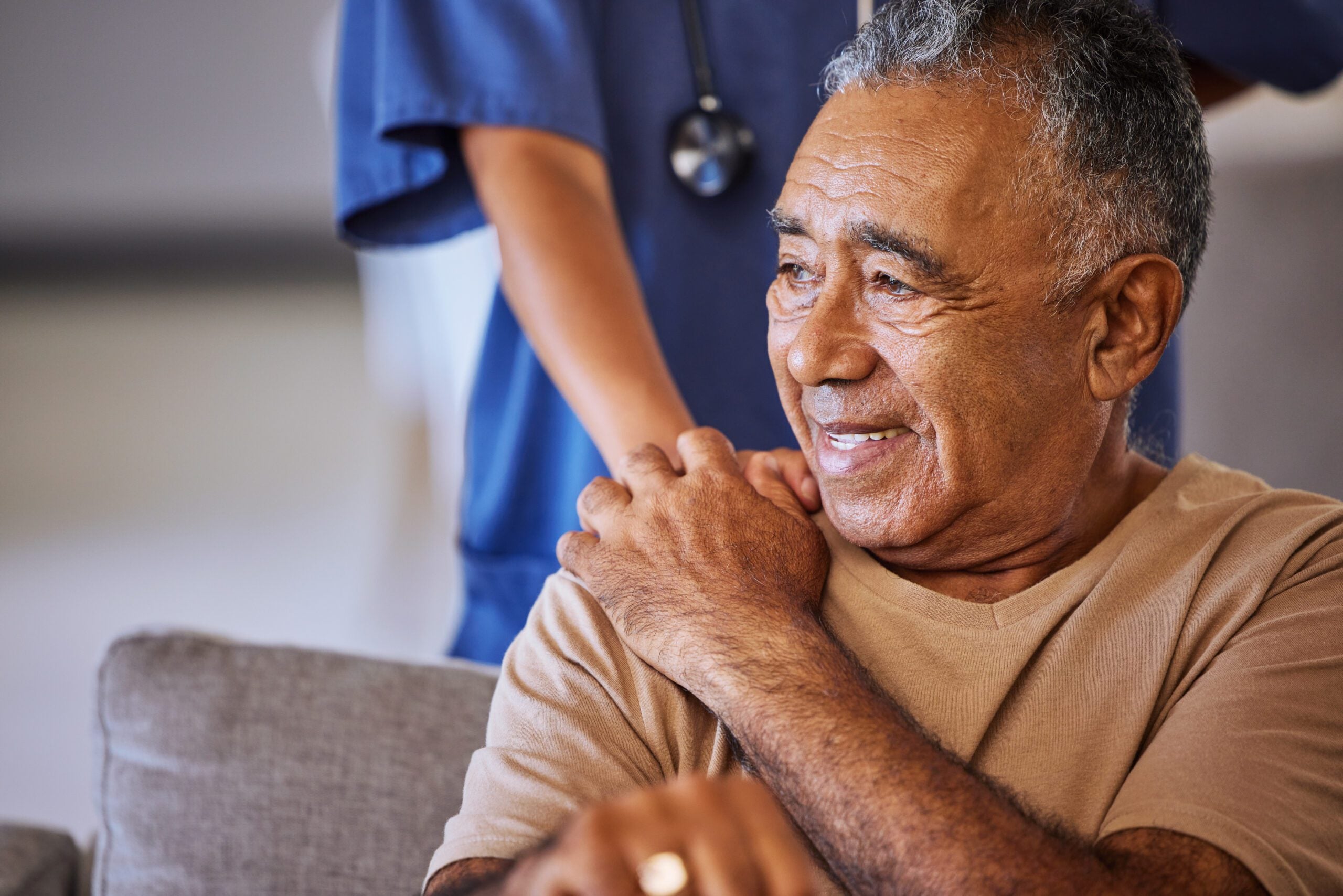 A man touches the hand of a care provider who is standing behind him with their hand on his shoulder.