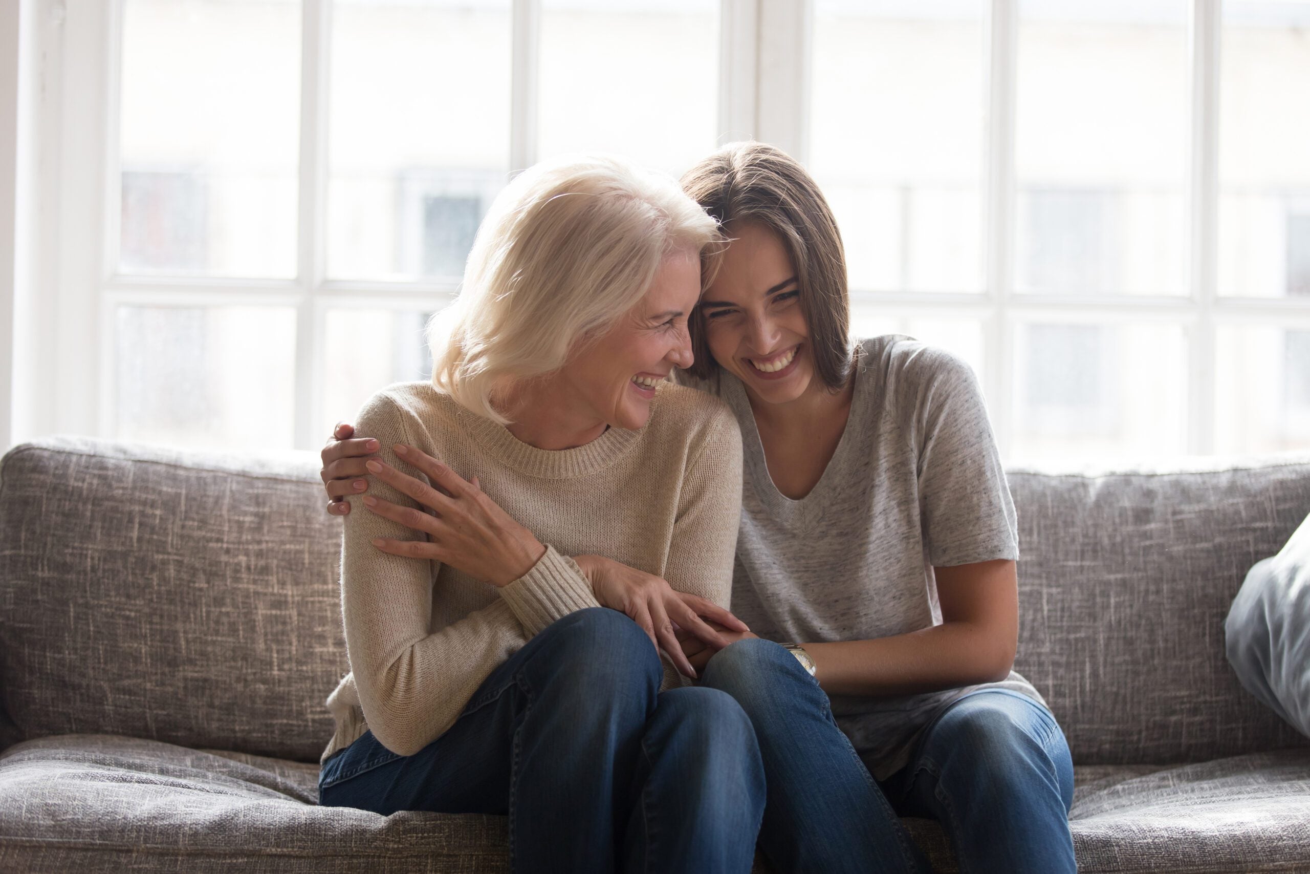 A woman and her senior mother sit on a couch, laughing together and embracing.