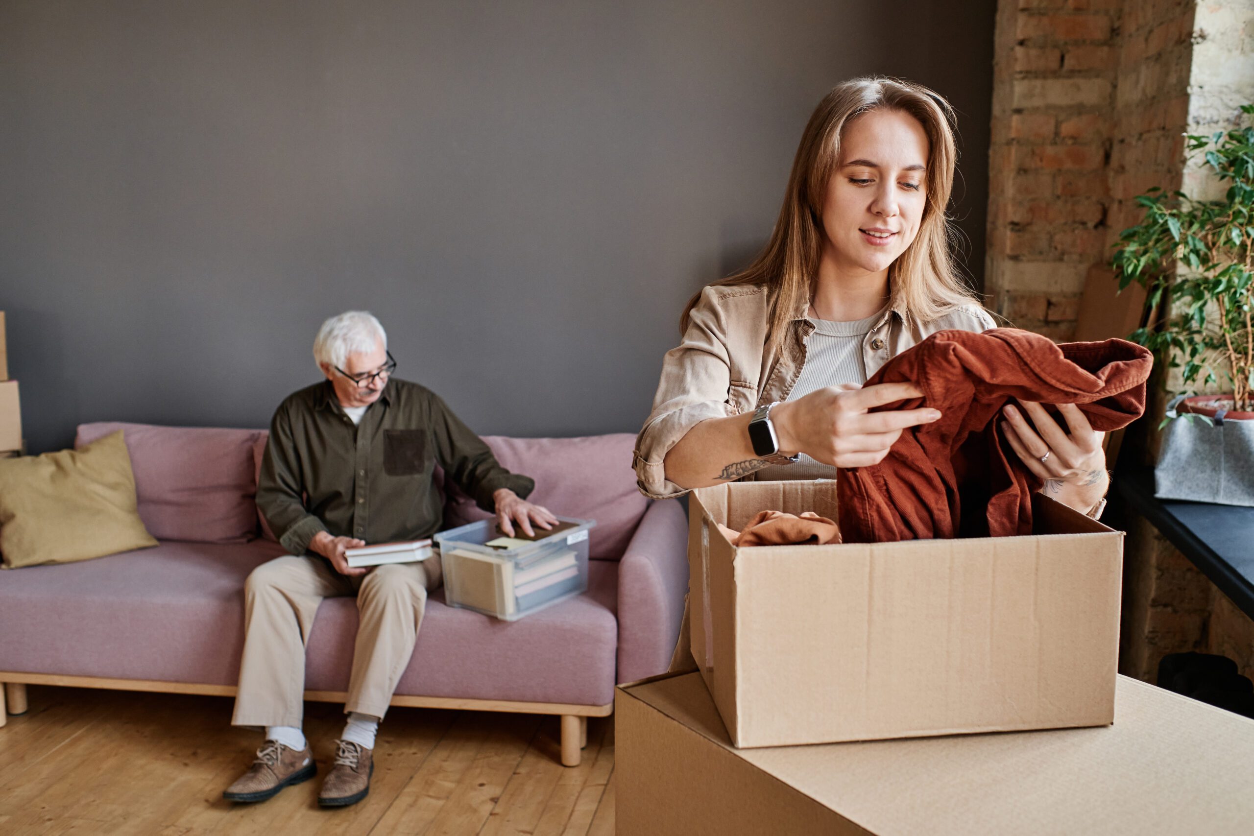 An adult child helps pack up their older parent’s belongings while the parents sit in the background sorting a box.