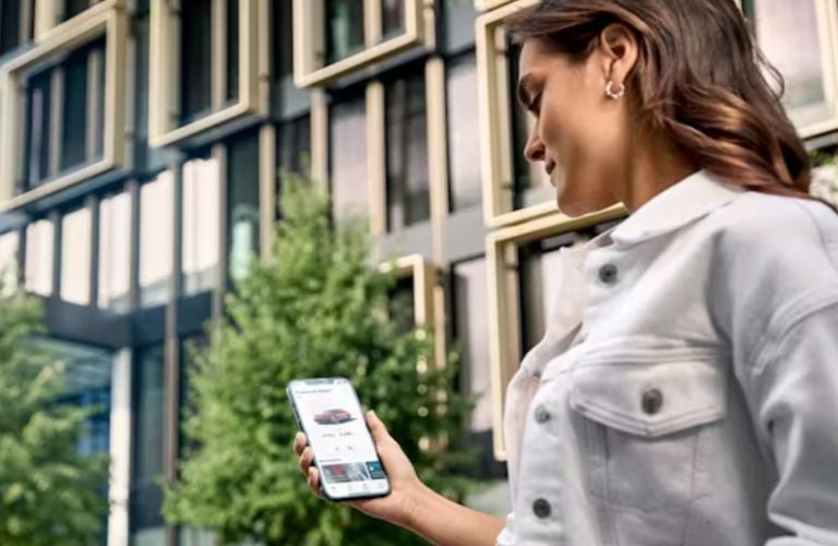 A woman in a white jacket holds a smartphone displaying a red car on a car-sharing app.