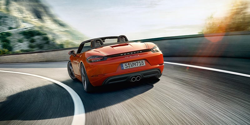 Rear view of a red Porsche 718 Boxster S convertible speeding right on a mountain road at sunset.