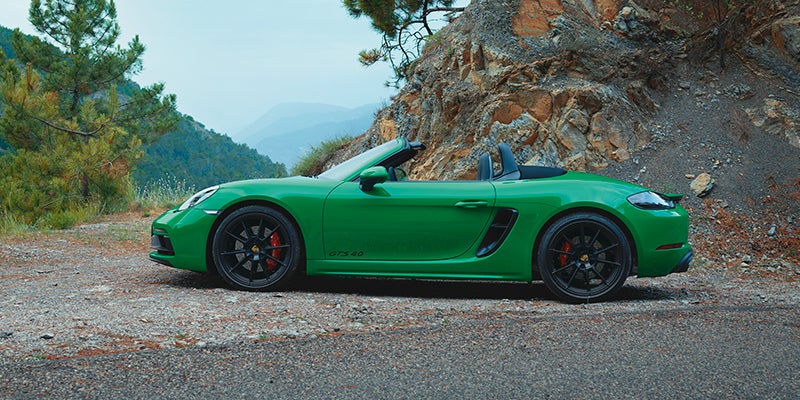 A bright green convertible sports car with an open top parked on a dirt road, mountains in the background.