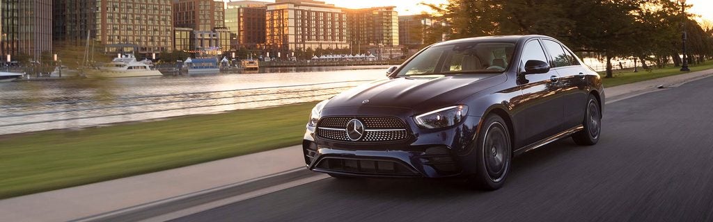 Black Mercedes-Benz E-Class sedan driving along a waterfront at sunset.