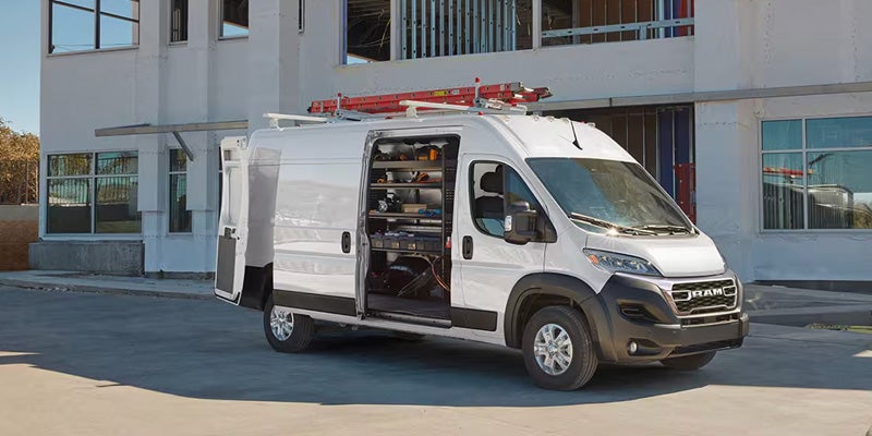 White RAM ProMaster cargo van with open side and rear doors, showing interior shelving and tools, outside a building.