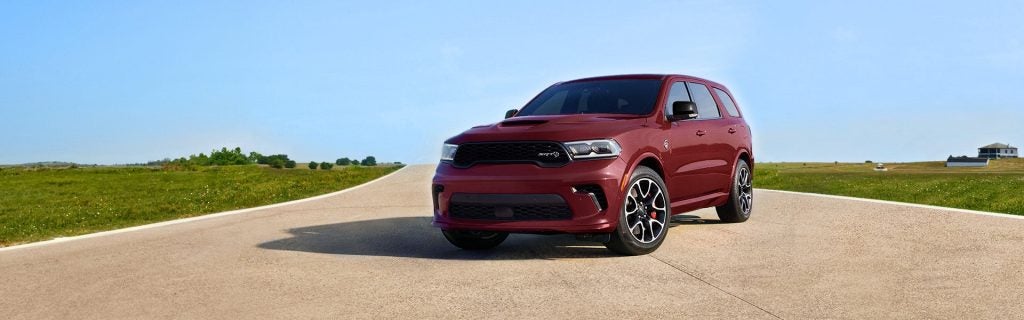 A red Dodge Durango SUV parked on a winding asphalt road, surrounded by green fields under a clear blue sky.