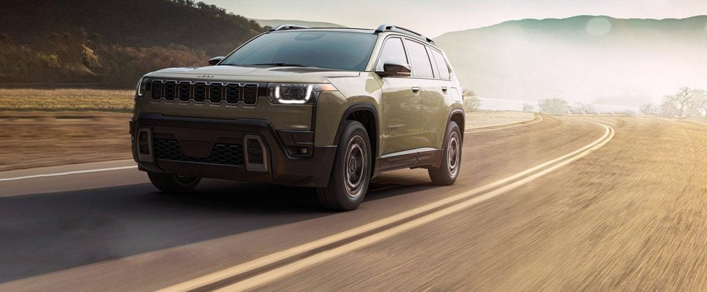 A light tan Jeep Grand Cherokee driving on a winding road with a blurred landscape in the background.
