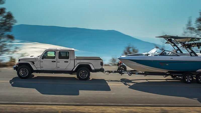 A white pickup truck towing a boat on a trailer down a road by a lake with mountains in the background.