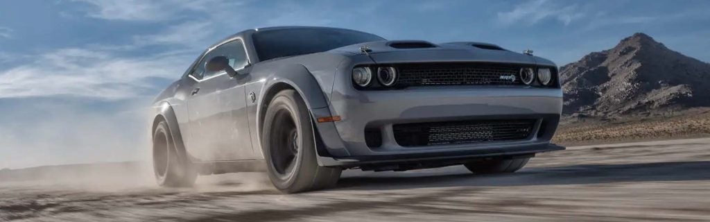 Silver Dodge Challenger car kicking up dust while driving right-to-left on a dirt road, with mountains in the background.