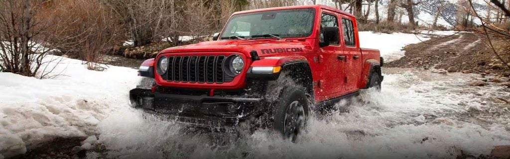 A red Jeep Gladiator pickup truck drives through a flowing stream, splashing water on snowy, wooded banks.