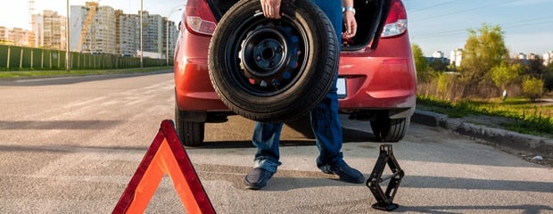 Person retrieving a spare car tire from a red car trunk, with a safety triangle and jack on the road.
