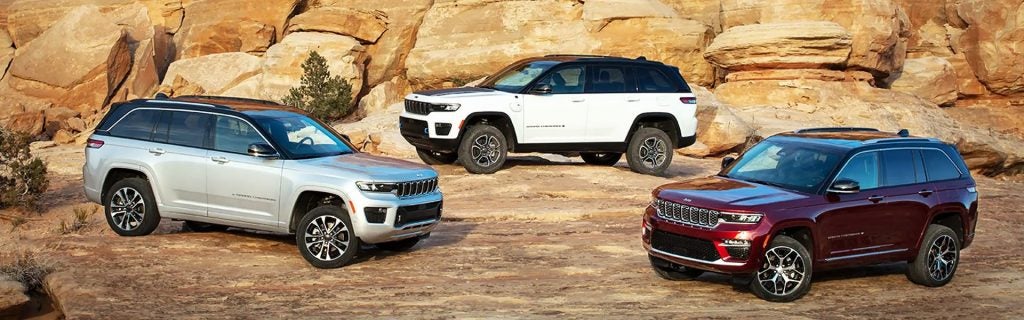 Three Jeep Grand Cherokee SUVs—silver, white, and burgundy—parked on a rocky, desert landscape.