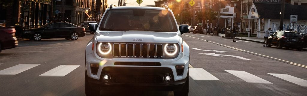 Front of a white Jeep Renegade driving down a city street at sunset, with buildings and other cars visible.