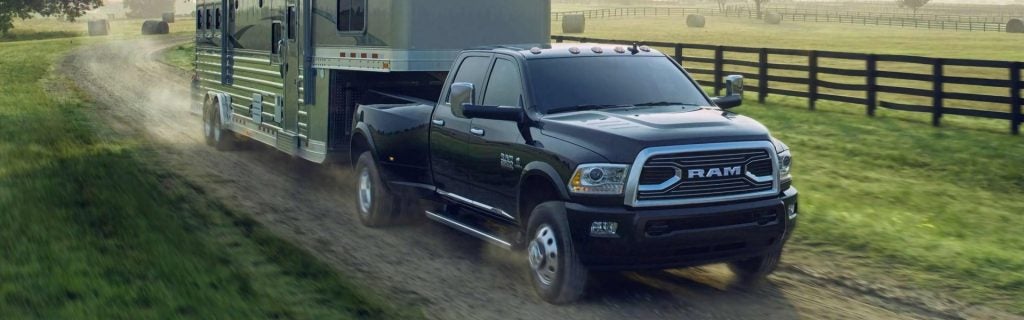 A black Ram dually truck pulling a large silver horse trailer on a dusty winding dirt road through green fields.
