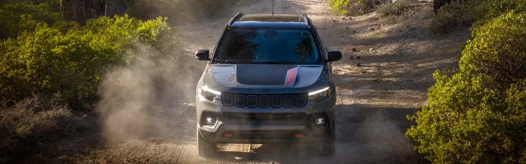 A dark grey Jeep SUV drives on a dusty dirt road, kicked-up dust visible, surrounded by green shrubs.
