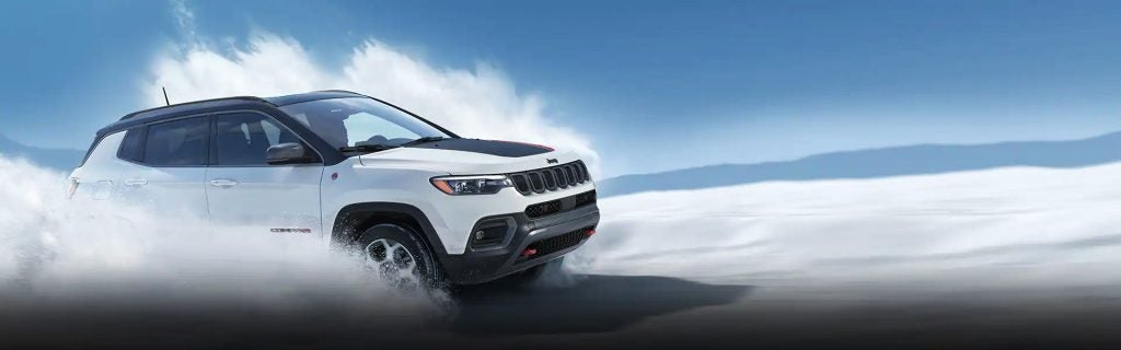 White Jeep Compass driving through water or snow, kicking up spray under a blue sky.